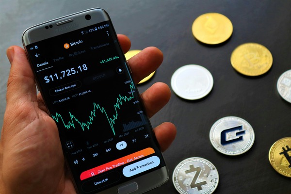 Hand holding a phone showing a Bitcoin trading app next to various cryptocurrency coins