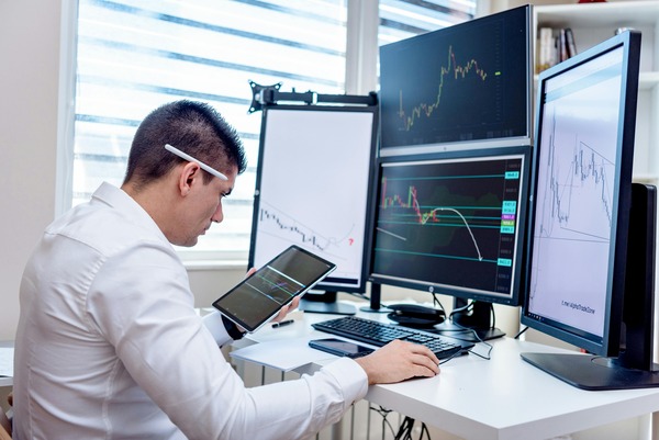 Trader reviewing chart data on a tablet next to multiple monitors